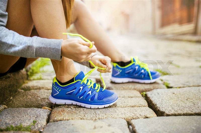Female athlete tying sport shoes laces for running on tiled pavment in city center. Runner getting ready for training, stock photo