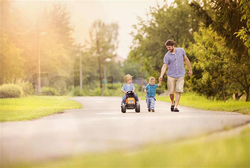 Happy young father with his two little sons have fun on road, one boy is riding on little tractor, other boy is holding father’s hand, stock photo