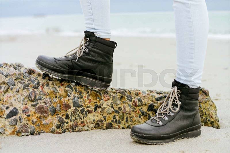 Female feet in blue jeans and black winter boots standing on a stone against the sea, stock photo