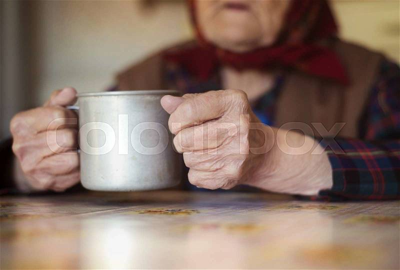 Very old woman in head scarf is drinking tea in her old country style kitchen, stock photo