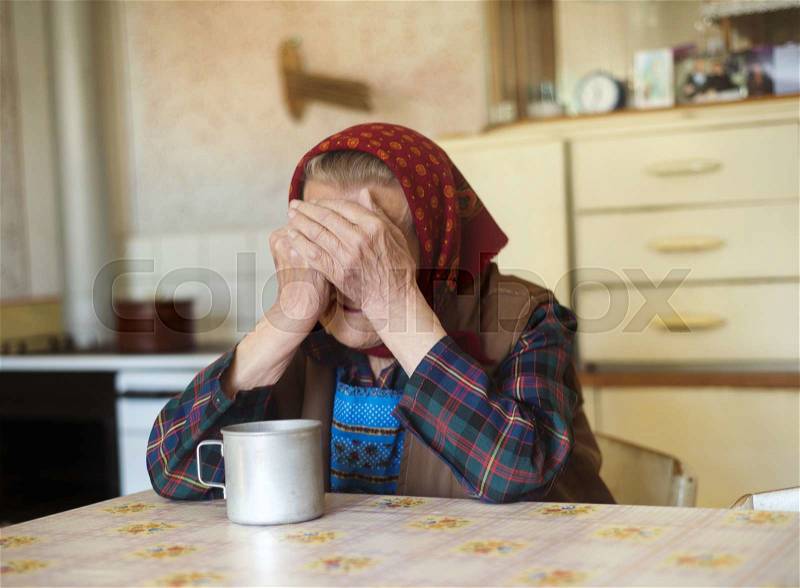 Very old sad woman in headscarf sitting in her countrystyle kitchen, stock photo