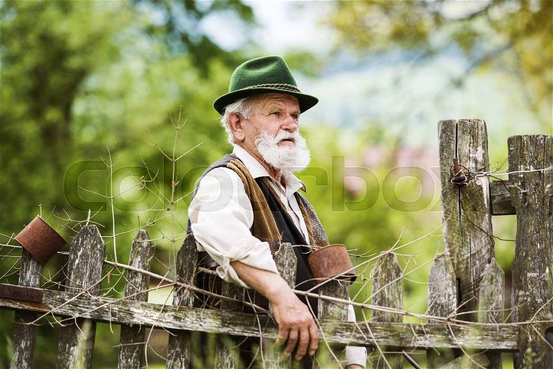 Old farmer with beard and hat standing by the lath fence with empty tins on top, stock photo