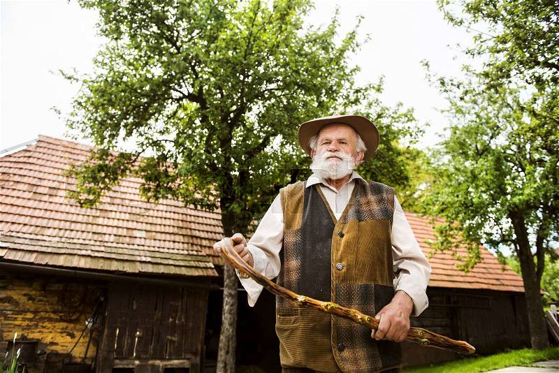 Old farmer with beard and hat standing by his farmhouse, stock photo