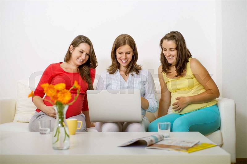 Three pregnant women sitting on sofa, chatting and using laptop, stock photo