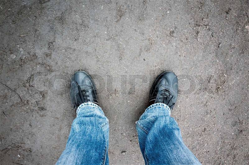Male feet in blue jeans and black shoes standing on dirty rural road, first person view , stock photo
