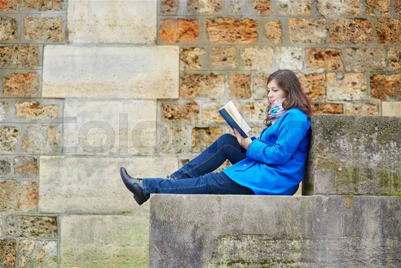 Beautiful young girl reading a book on the Seine embankment in Paris, stock photo