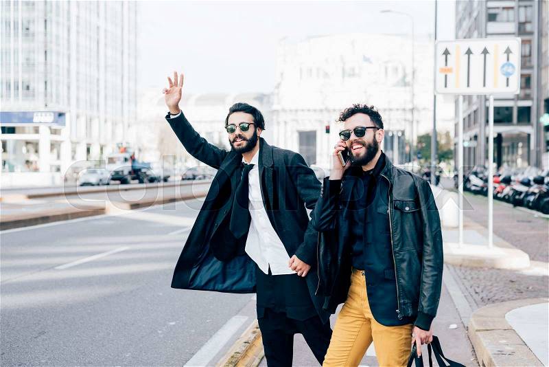 Knee figure of two young handsome caucasian bearded brown hair businessmen outdoor in the city, one asking for a taxi, the other talking smartrphone - business, technology, communication concept, stock photo
