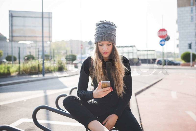 Knee figure of young handsome caucasian blonde straight hair woman sitting outdoor in the city, smartphone handhold, looking down tapping the screen - communication, technology, social network concept, stock photo