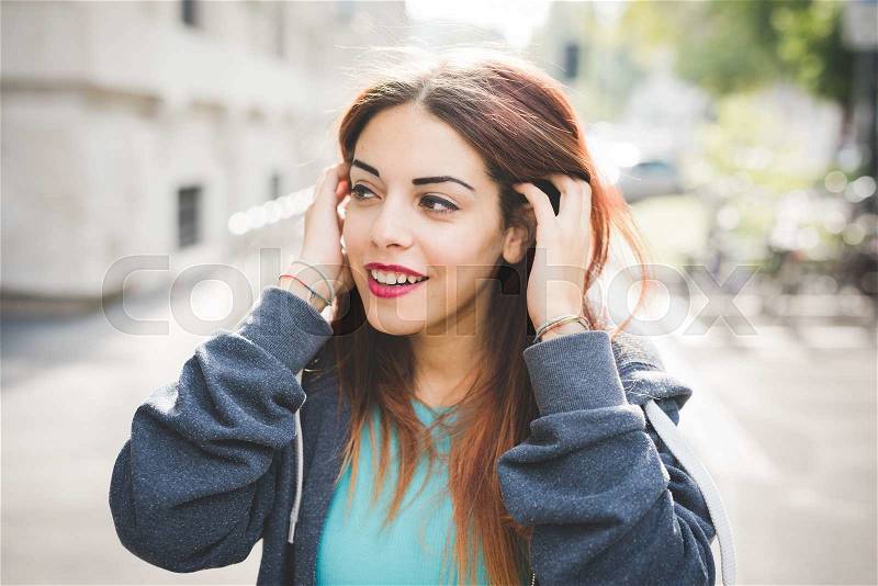Half length of young caucasian reddish hair woman outdoor in the city brushing her hair behind her ears, overlooking right, smiling - wearing blue sweatshirt and azure shirt - youth, carefree concept, stock photo