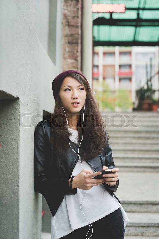 Knee figure of young handsome asiatic chinese woman posing leaning against a wall, listening music with earphones and smartphone handhold, overlooking left - music, technology concept, stock photo