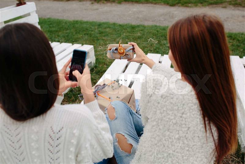 Two girls sit on a bench outside and shoot gifts for smartphone, stock photo