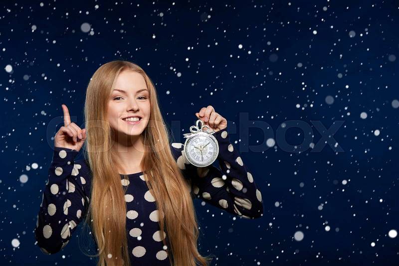 Christmas, New Year, winter holidays celebration concept. Smiling woman holding a clock over snow background and pointing her finger up, stock photo