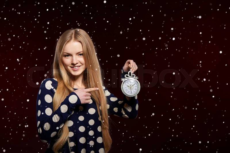 Christmas, New Year, winter holidays celebration concept. Smiling woman holding a clock over snowy background and pointing at it, stock photo