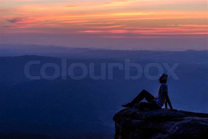 Man sitting on mountain peak at sunset with mountains background, stock photo