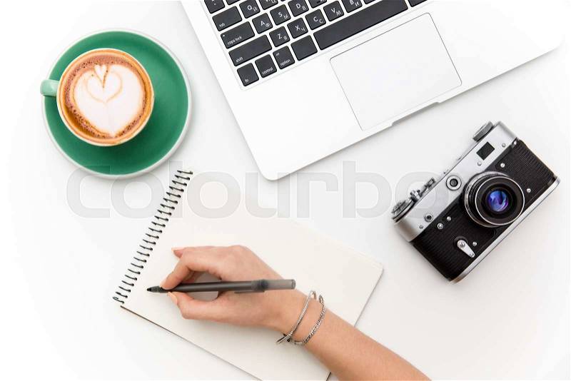 Top view of laptop, old camera, cup of coffee and woman hand writing in notebook over white background, stock photo
