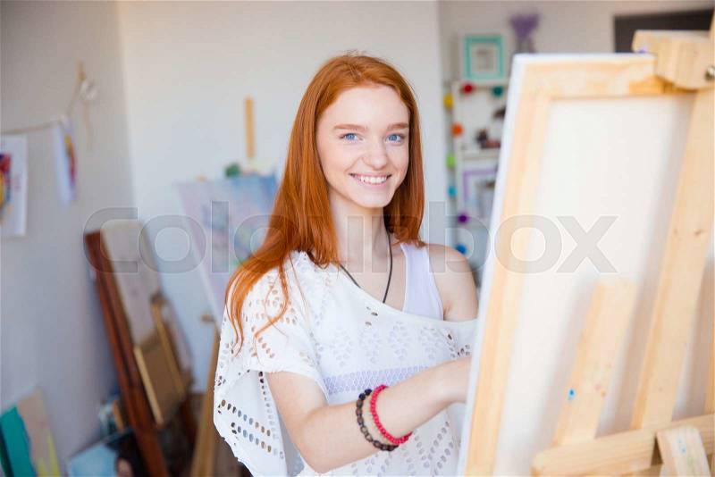 Cheerful attractive woman artist painting on canvas in art workshop, stock photo