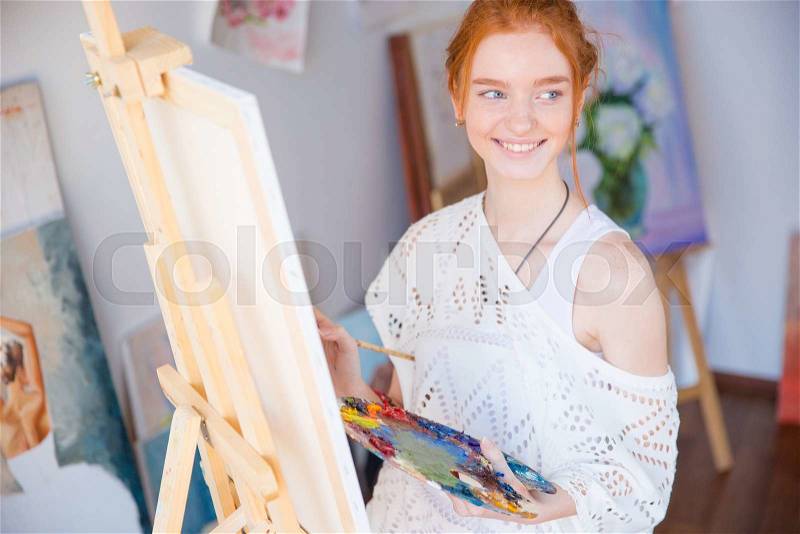 Happy lovely cute young woman painter holding palette with oil paints and painting on canvas in art studio, stock photo