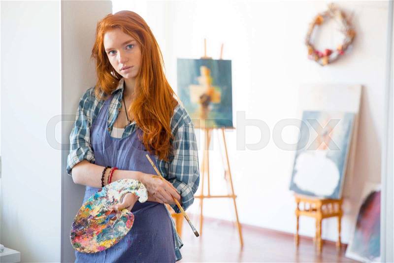 Thoughtful atractive young woman painter with long red hair in apron holding art palette and brush in artist workshop, stock photo