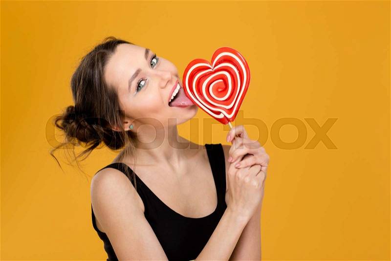 Lovely playful young woman licking sweet heart shaped lollipop over yellow background, stock photo
