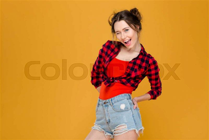 Playful happy young woman in plaid shirt and jeans shorts standing and winking , stock photo