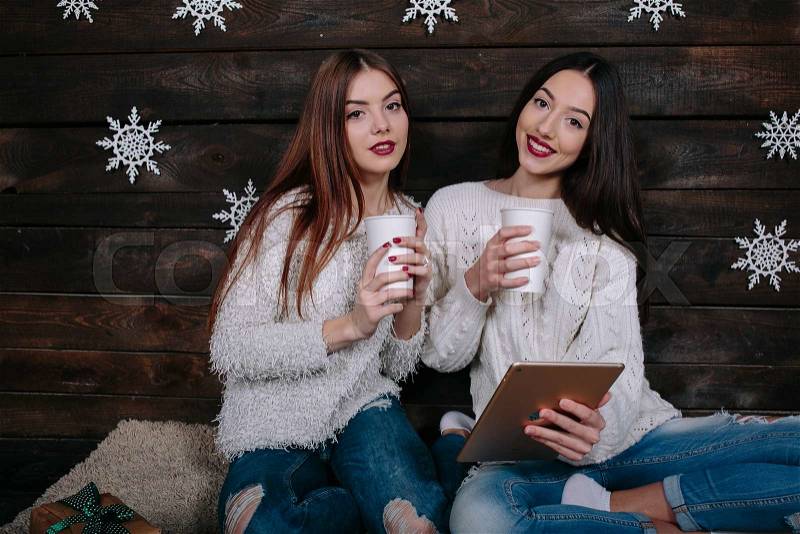 Two girls drink a hot drink and watching something on a tablet, stock photo