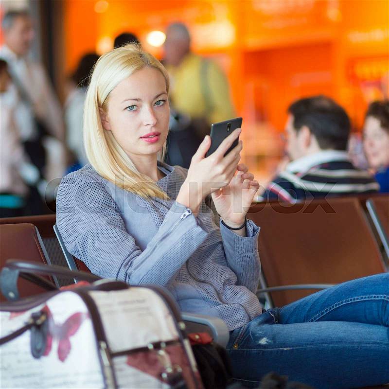 Casual blond young woman using her cell phone while waiting to board a plane at the departure gates. Wireless network hotspot enabling people to access internet conection. Public transport, stock photo