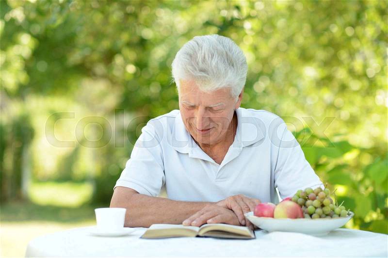 Handsome older man sitting at a table at home on the veranda with book, stock photo