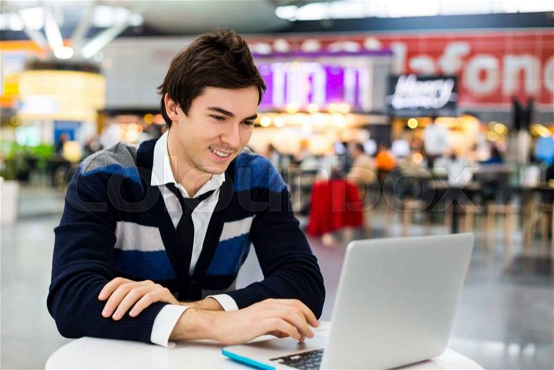 Happy young business man work on computer outdoor on white table. Guy is freelancer and handsome and look at laptop. Freelancer workspace at the airport or train station, stock photo