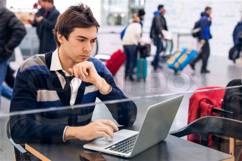 Freelancer working with a laptop and phone in a train station while is waiting for transport. On a background people walking with suitcases and bags. Busy guy sitting at the airport or train station, stock photo