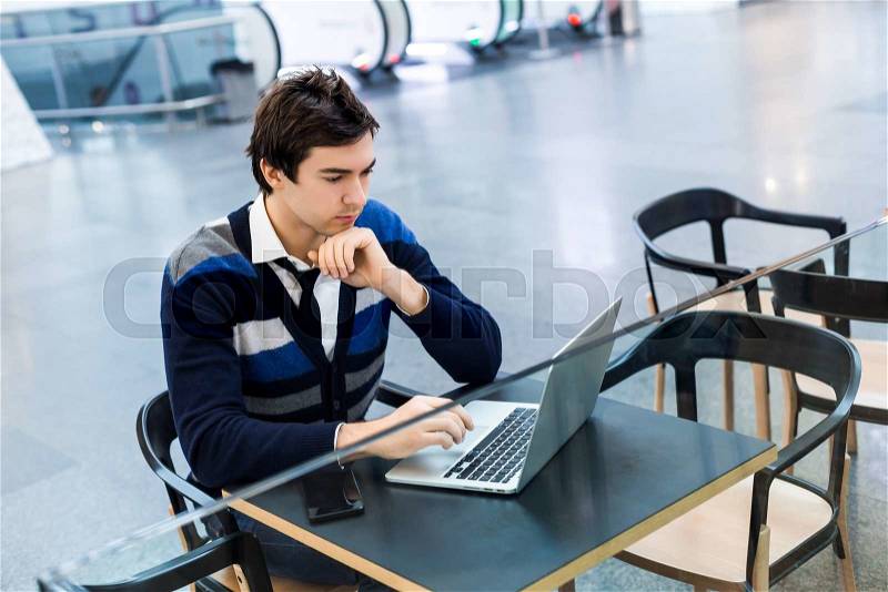 Entrepreneur man working with laptop in restaurant or in airport cafe. Busy guy freelancer guy sitting with open computer and concentrated work. On a background people walking with suitcases and bags, stock photo