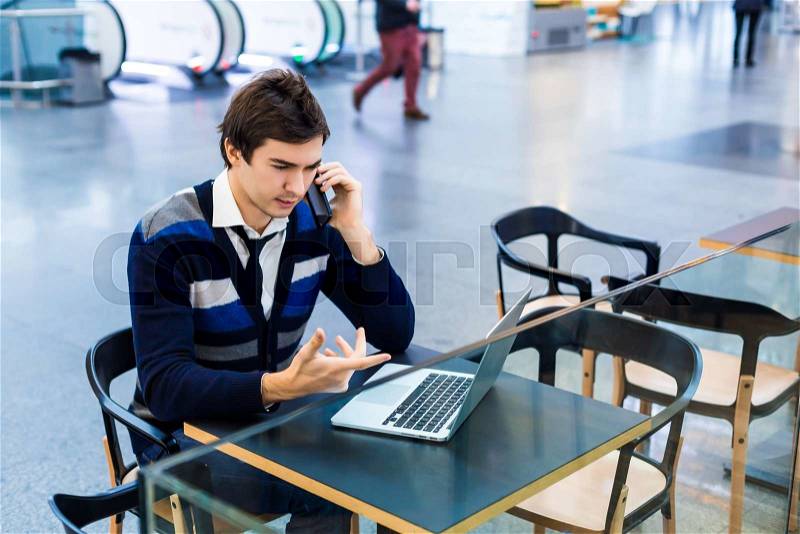 Young freelance man using mobile phone and laptop pc.Teenager student talking on smartphone in airport cafe. Man serious and hand gestures.Freelancer workspace at the airport or train station, stock photo