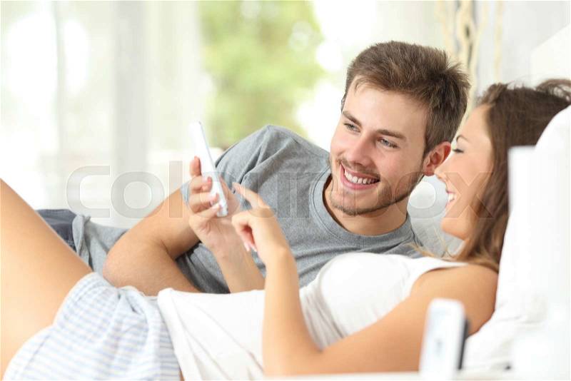 Happy marriage sharing a smart phone on the bed at home, stock photo