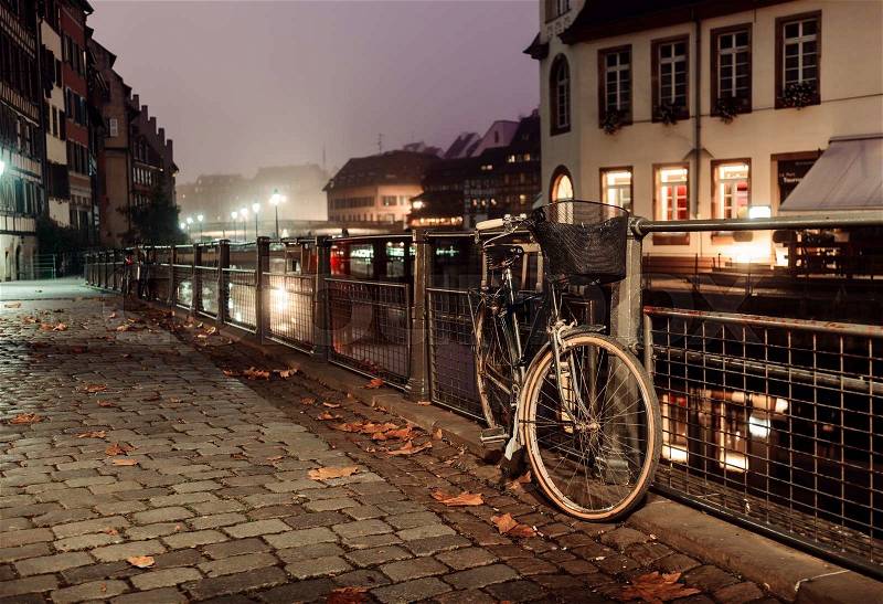 Old bicycle on city cobblestones fastened to the fence that runs along the river lit by the lights of the city at night, stock photo