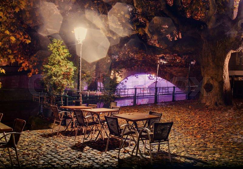 Empty tables under a lamp at night in fog on the city promenade in autumn fallen leaves on a cobblestone street in the background Bridge over the river, stock photo
