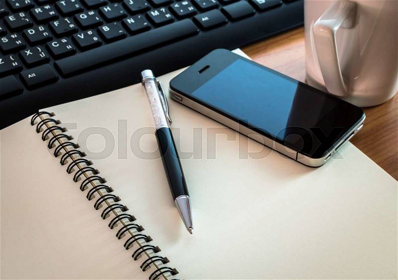 Pen on notebook with computer keyboard, coffee cup and cell phone on wooden table, stock photo