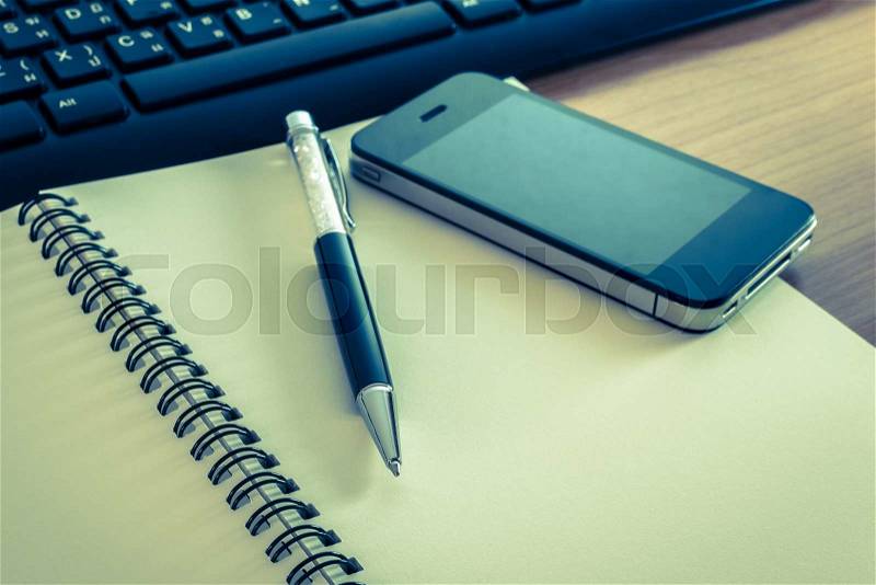 Pen and cell phone on a notebook with keyboard on wooden table in cross processed color, stock photo