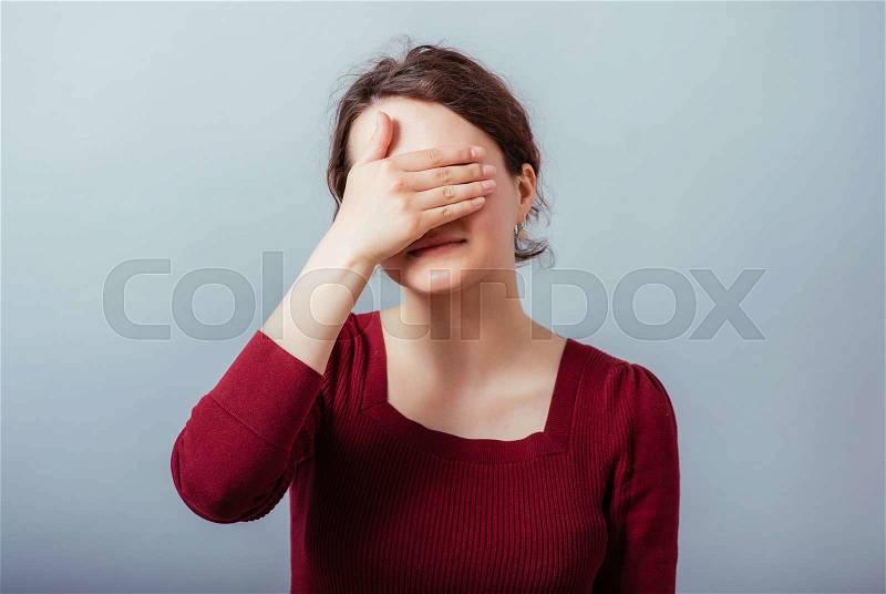 Young sad woman covering her eyes with her hand on the eyes. On a gray background, stock photo