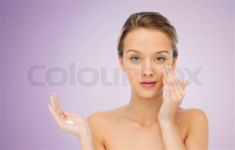 Beauty, people, cosmetics, skincare and health concept - young woman applying cream to her face over violet background, stock photo