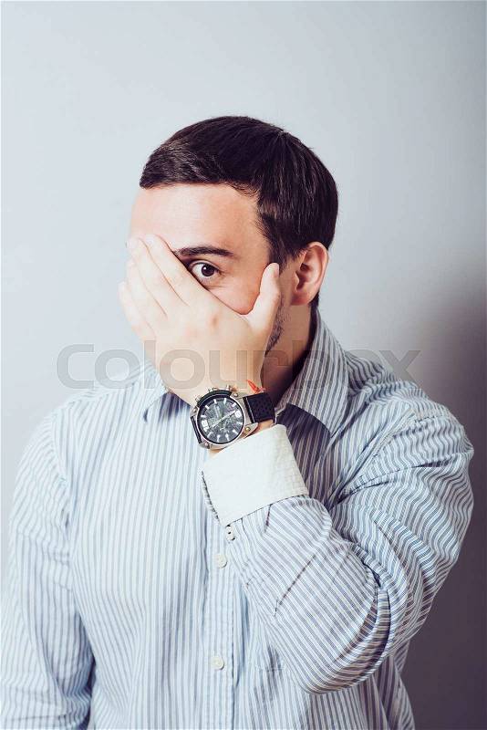 Shocked and terrified. Portrait of young man covering his face by hand and looking at camera while standing against grey background, stock photo