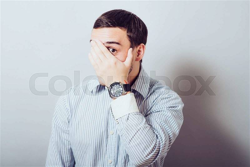 Shocked and terrified. Portrait of young man covering his face by hand and looking at camera while standing against grey background, stock photo