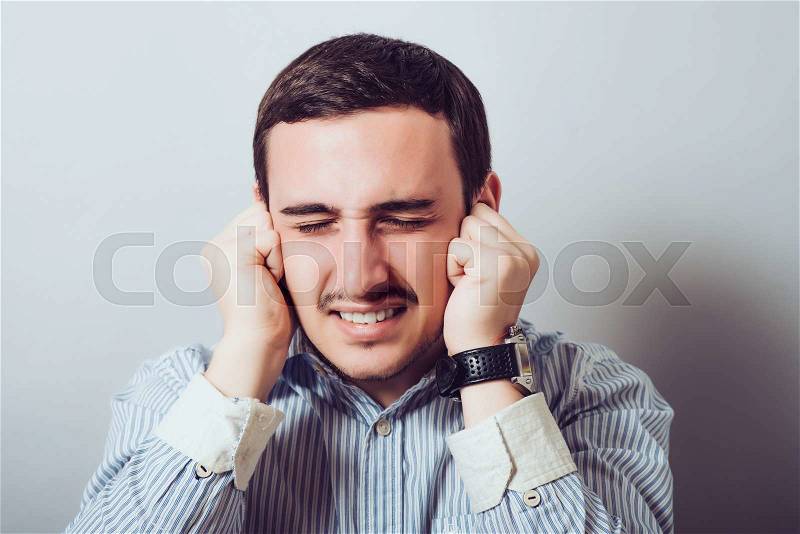 This is too loud! Frustrated young man holding head in hands and keeping eyes closed while standing against a gray background, stock photo