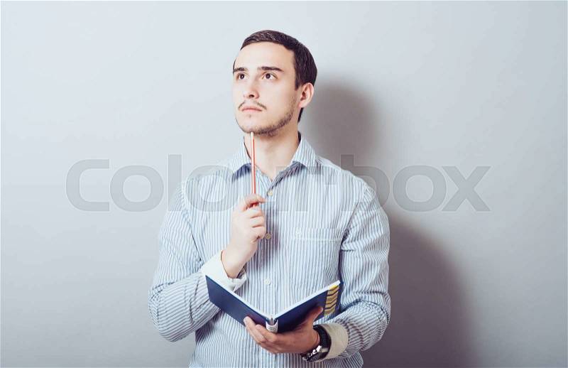 Young man is planning something, making notes with a pencil and notepad. On a gray background, stock photo