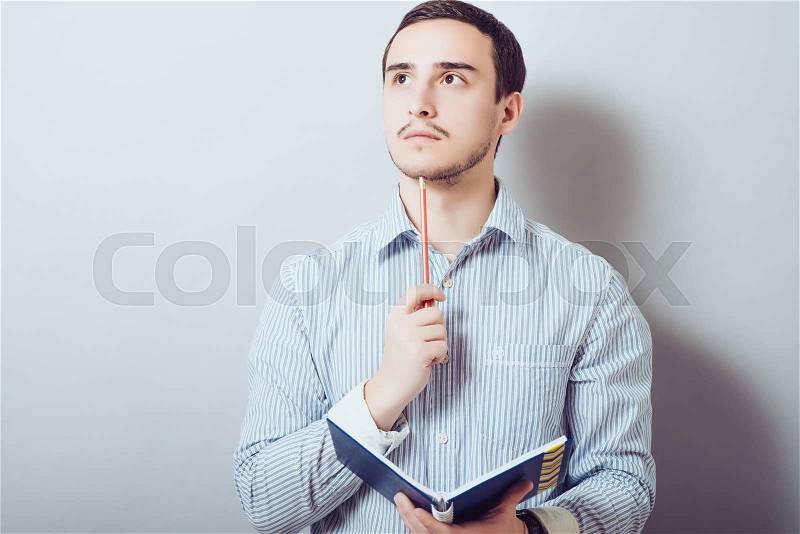 Young man is planning something, making notes with a pencil and notepad. On a gray background, stock photo