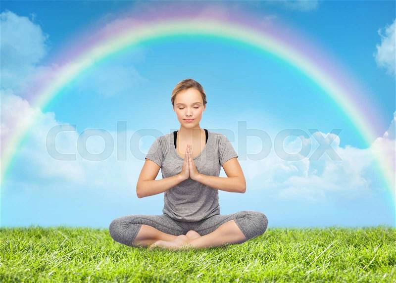 Fitness, people, harmony and healthy lifestyle concept - woman making yoga meditation in lotus pose over grass and rainbow in blue sky background, stock photo