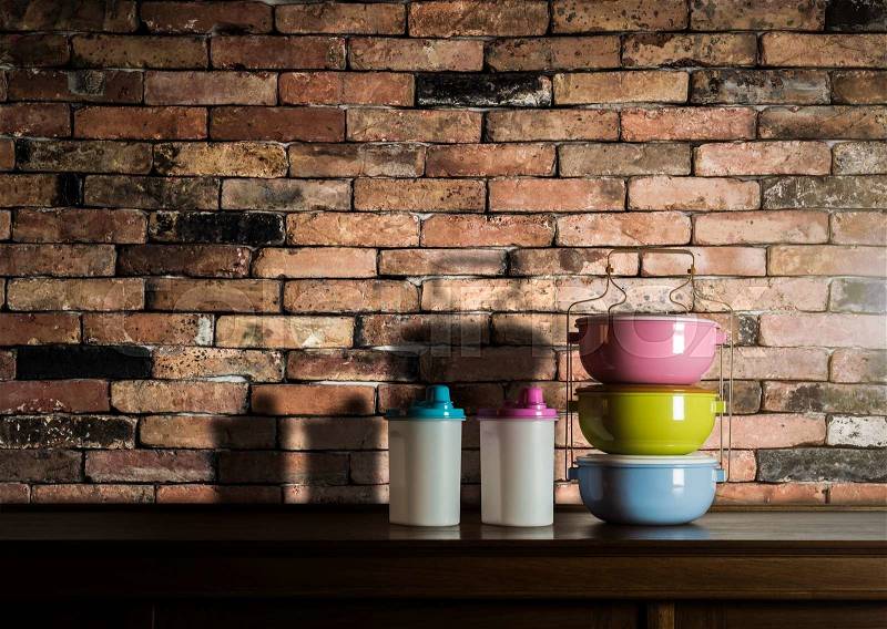 Colorful tiffin carrier and plastic bottles on wooden cupboard with vintage brick wall background against warm light, stock photo