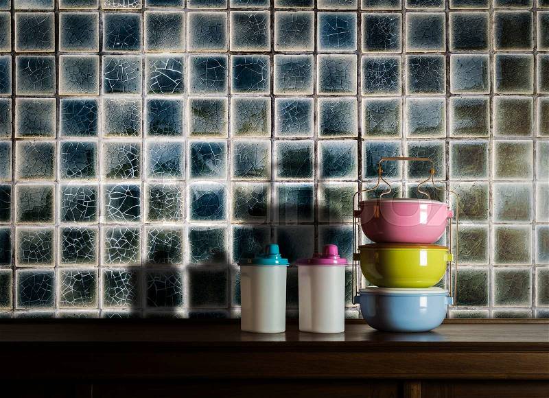 Colorful tiffin carrier and plastic bottles on wooden cupboard with vintage tiles wall background against warm light, stock photo