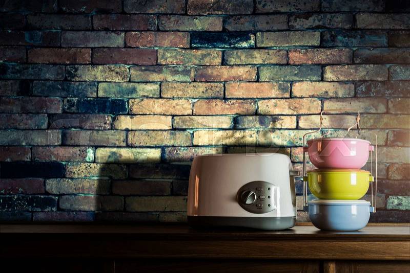 Colorful tiffin carrier and toaster on wooden cupboard with vintage brick wall background against warm light, stock photo