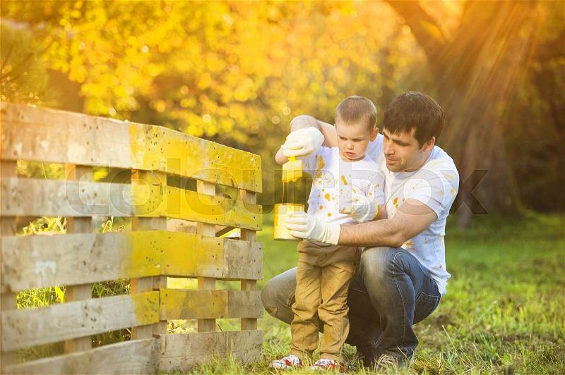 Cute little boy and his father painting wooden fence together on sunny day in nature, stock photo
