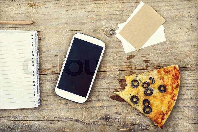Desktop mix with office gadgets, supplies and pizza on a wooden office table background. View from above, stock photo