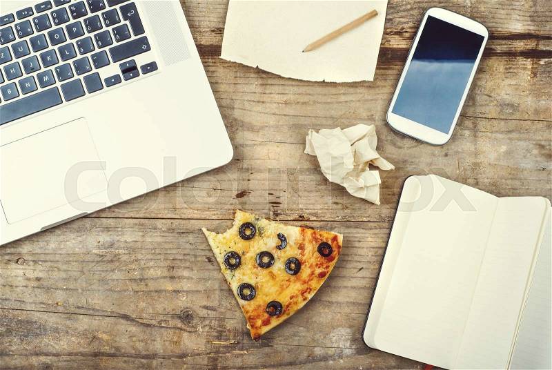 Desktop mix with office gadgets, supplies and pizza on a wooden office table background. View from above, stock photo